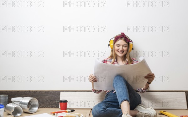 Happy girl in protective headphones sitting on floor with paper blueprint. Home remodeling after moving. Construction tools and materials for building. Young woman wearing checkered shirt and jeans