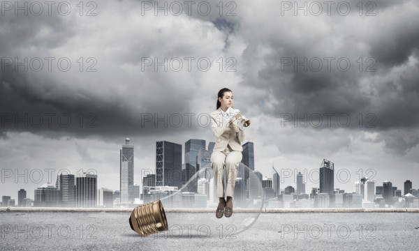 Attractive woman playing trumpet brass on background modern city and storming sky. Young businesslady in white suit sitting on big light bulb with music instrument. Musician practicing and performing