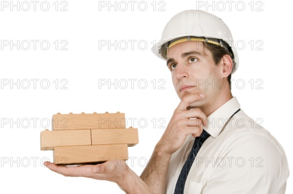 Upper body shot of a young man in a white shirt and white helmet with a stacked wooden block on his left hand and with his right hand on his chin looking upwards in front of a white background