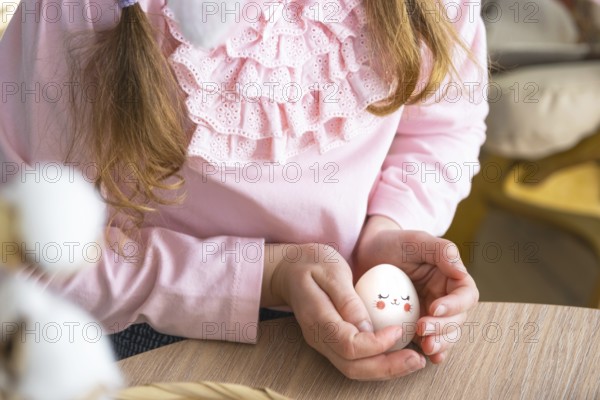 A cute girl with pink bunny ears makes an Easter craft, decorates an egg in the form of a unicorn with rhinestones, horn, flowers in the interior of a house with plants