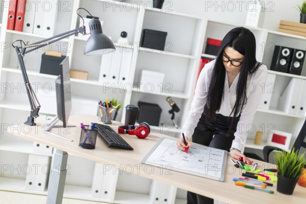 A young girl with glasses, wearing a white blouse and black trousers, works in the office. The girl has long straight black hair. photo with depth of field