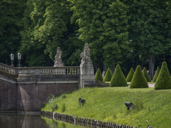 A stone bridge with statues leads over a river, flanked by neat hedges and lush trees, nordkirchen, münsterland, germany