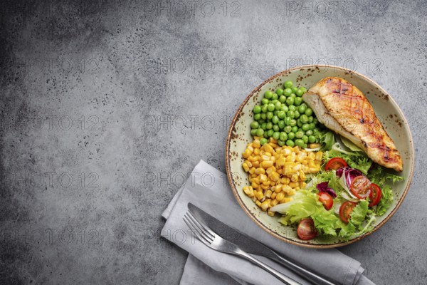 Baked chicken breast on plate with fresh salad, green peas and corn, grey stone background, top view. Healthy fitness meal with chicken fillet, balanced in proteins and carbs. Overhead, copy space