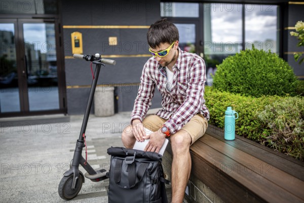 Handsome hipster style bearded man works online freelancer, pulls out laptop with backpack and electric scooter, active lifestyle, backpacker traveller. Ecology and clean ecological mode of transport
