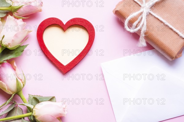 Valentine day theme image with a red heart with copy space in the middle, surrounded by pink roses, an envelope and a gift wrapped in brown paper, on a pink background