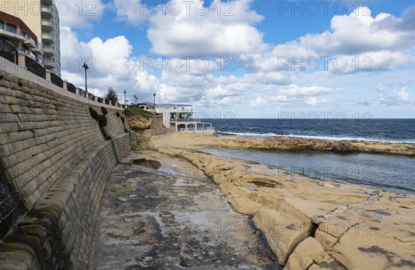 Saint Julian, Malta, 01 07 2022: Low angle view over the water, rocks and sand at the beach with the harbor in the background