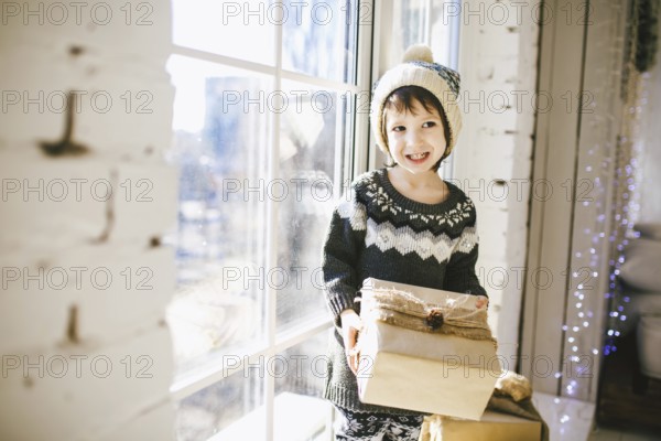 Child sitting at the window on a sunny Christmas day, making presents wrapped in boxes in paper. dressed in knitted warm woollen clothes and hat. Inside the house with a stylish interior