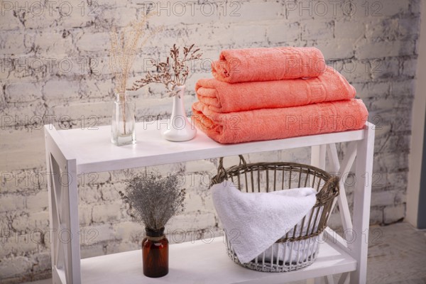 Rack with a stack of three peach colour towels and baskets with clean white towels and toilet decor near a brick wall. Shelf with terry towels and decorative vases. Shelving unit with clean towels