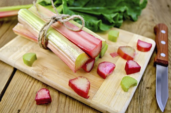 Bundle of stalks rhubarb, cut pieces of rhubarb with a sheet and a knife on a wooden boards background