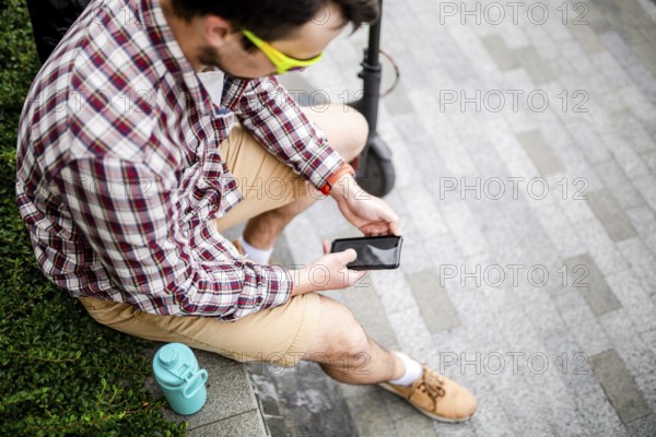 Caucasian man uses modern technologies Internet and cellular communication on smartphone while sitting on bench in city near electric scooter. Ecology and environmentally friendly urban transport
