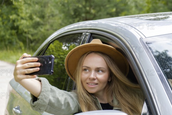 Exited smiling woman making video call with mobile phone from car window. Local solo travel on weekends concept. Young traveler explore freedom outdoors in forest taking selfie photo. Unity with nature lifestyle, rest recharge relaxation