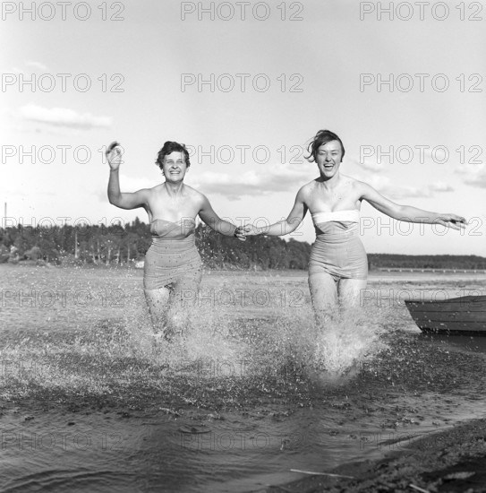 1950s summer. Two young women runs on the shallow part of the beach and the water splashes. Sweden 1959. Kristoffersson ref CG10-8