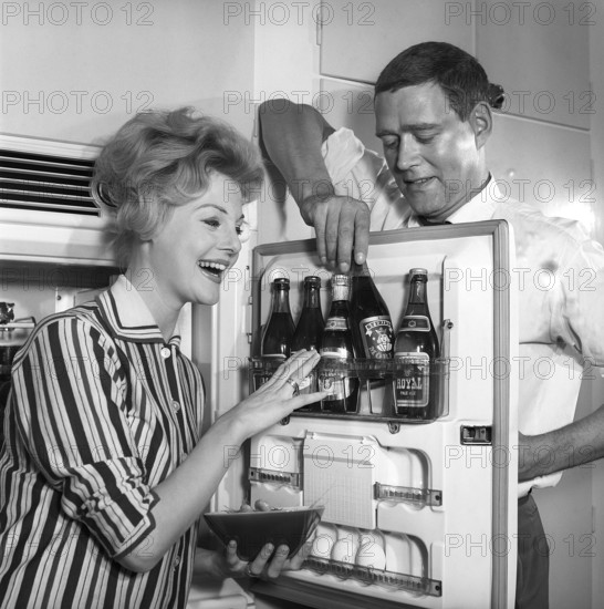 In the kitchen 1950s. A couple in their kitchen and at the refrigerator where the beer is kept in the door shelf. Five beer bottles are seen. Mrs is holding a bowl of shrimps. Sweden 1959. Kristoffersson ref CH77-3