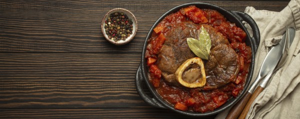 Traditional Italian dish Ossobuco all Milanese made with cut veal shank meat with vegetable tomato sauce served in black casserole pan top view on rustic brown wooden background, copy space, food photography, food photography