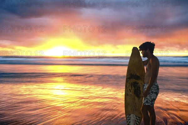A surfer stands with surfboard in hand on the beach and watches the sun rise over the ocean on a cloudy day, creating stunning colours in the clouds, on the ocean and on the beach