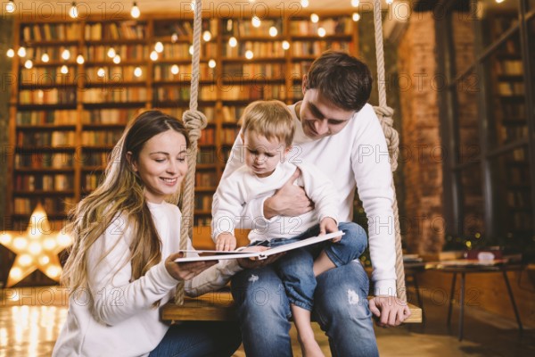 Family togetherness. Mom, dad and son reading story book together sitting on swing evening at home. Family and Parenthood Concept. Happy family reads childrens book while sitting home in loft interior