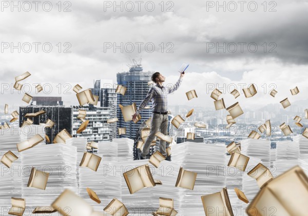 Man in casual wear keeping hand with book up while standing among flying books with cityscape on background. Mixed media