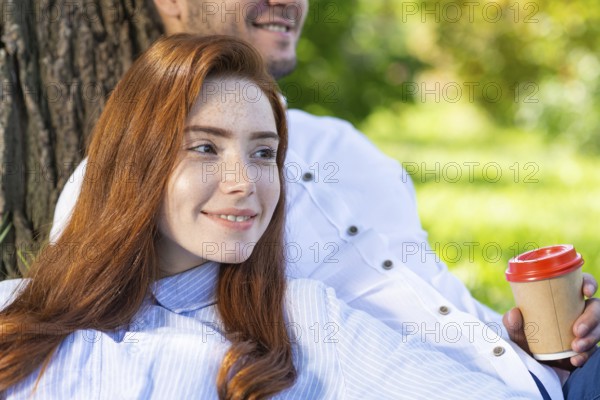 Young couple relaxing under tree in summer park on sunny day. Happy couple in love spend time outdoors together. Handsome man and pretty redhead girl enjoying each other. Romantic relationships