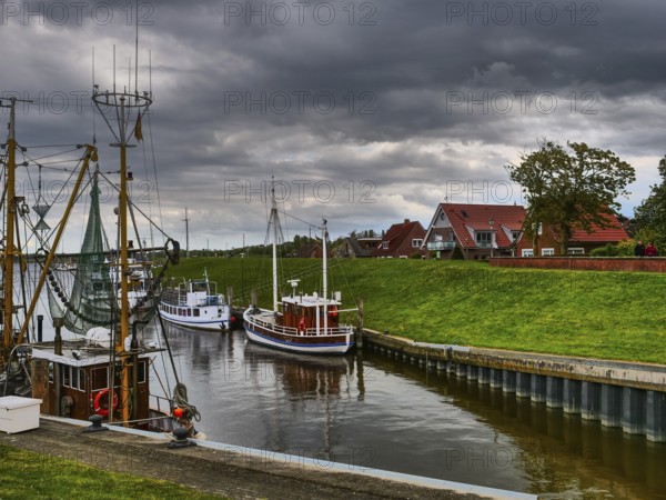 Boats in the river on a cloudy day, behind them houses and green embankment, Colourful fishing boats in a harbour on the North Sea with old houses in the background, Greetisel, Germany