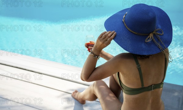 Woman in blue hat and glass of wine is relaxing near the pool