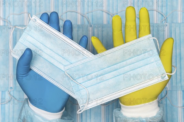 Doctor holds medical face mask in blue glove in one hand and other surgical face mask in yellow protective glove. Hygiene concept image on background of group of blue respiratory bandages for human