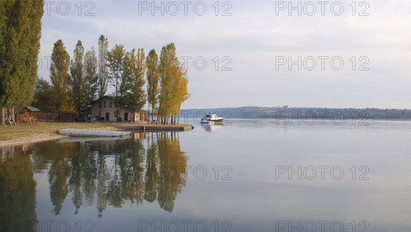 Scenic view with a house on the riverbank of Nistru river and a yacht floating. Autumn season landscape, colorful trees and beautiful sky reflected on the water surface