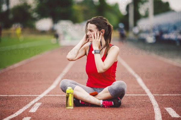 Theme healthy lifestyle, sports music. beautiful young Caucasian woman rests sitting treadmill running rubber stadium uses technodogy mobile phone hand insert earphones into ear include music training