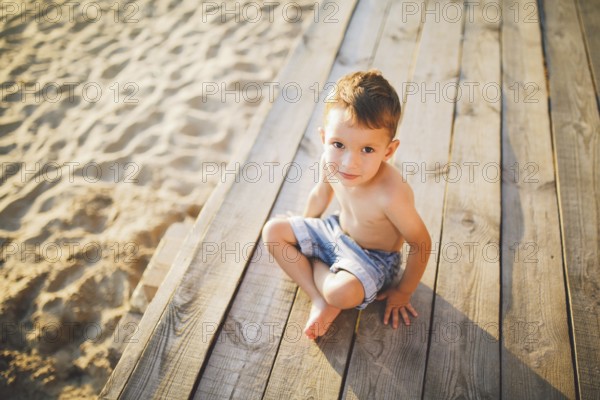 Little Caucasian boy child sitting on wooden pier sandy beach, summer time, sea holiday near water. The theme is the flow of time, a short life, the meaning and purpose of existence. Meaning of life