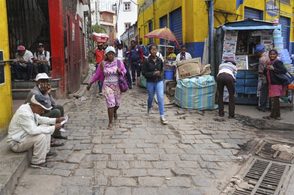 Antananarivo, Madagascar - April 24, 2019: Two unknown young Malagasy girls walking cobblestone road, more people around at their stalls, or sitting on ground - typical scene in streets of Tana city, Antananarivo, Madagascar