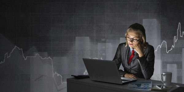 Attractive young businesswoman in glasses sitting at office desk and working at laptop. Female accountant in business suit and tie busy at work in evening. Financial analysis and planning
