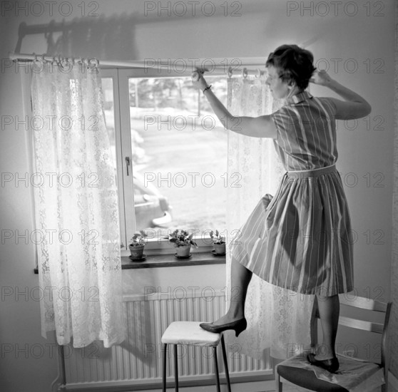 1960s home decorating. A woman at home putting up the curtains in the room window. All by herself she balances on a chair and a stool to manage it. Sweden 1966