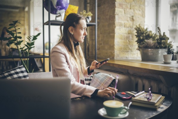 Woman sitting coffee shop wooden table, drinking coffee and using smartphone. Woman Using Phone In Cafe. Student learning online. Girl checking email. Woman Cafe, use mobile Phone, Working laptop
