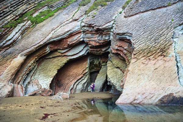 Huge cave on the beach. Low tide in the Atlantic. Flysch - unique landscape of underwater rocks exposed at low tide. Woman in pink jacket photographs Playa Zumaya