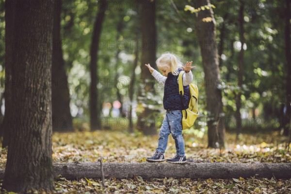 The theme children outdoor activities. Funny little baby Caucasian blond girl walks through forest overcoming obstacles, tree fell, log. Baby hiking big funny backpack in autumn forest park