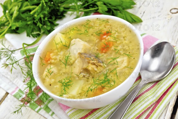 Soup fish kulesh with millet, potatoes and carrots in a white bowl on a napkin, parsley, dill on a wooden board background