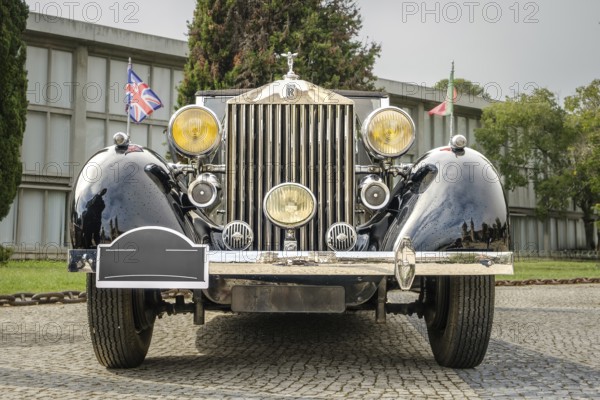 Lisbon, Portugal - Oct 15, 2023: Front view of Rolls-Royce Phantom III parked in cobblestone driveway. Full-size luxury car, 1930s, by Rolls-Royce Ltd, Britain, UK. Portuguese and British flags, hood mascot, Spirit of Ecstasy, grille, big headlights
