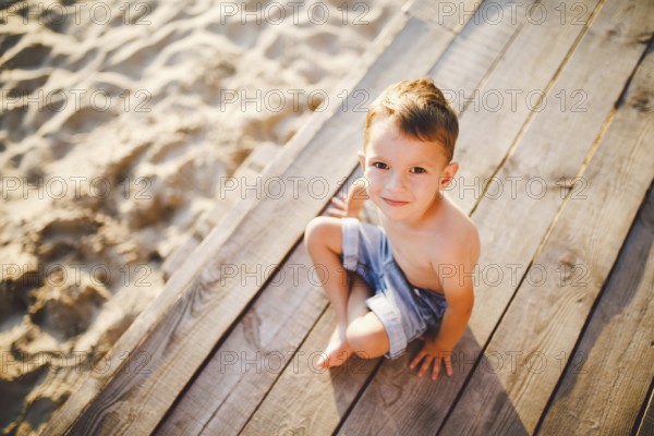 The theme is a child and summer beach holiday. A small Caucasian boy sits sideways on a wooden pier and looks at the camera on a sandy beach and a pond, a river. With bare legs in blue denim shorts