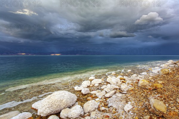 Rocky beach covered with evaporated salt. The Dead Sea is a closed salt lake. Gloomy sky with dark thunderclouds. Exotic resort for treatment and relaxation. Israeli coast