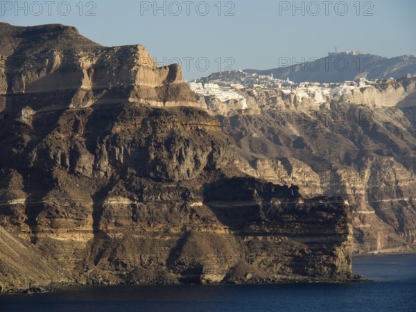 A wide mountain panorama with a sea and rocks in the foreground and a village in the background, brown volcanic island in the blue sea with white houses, Santorini, Greece