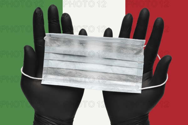 Doctor holds face mask in hands in black medical gloves on background colors flag of Italy or Italian Tricolour. Pandemic insurance coronavirus, flu, airborne diseases. Human medical respiratory mask