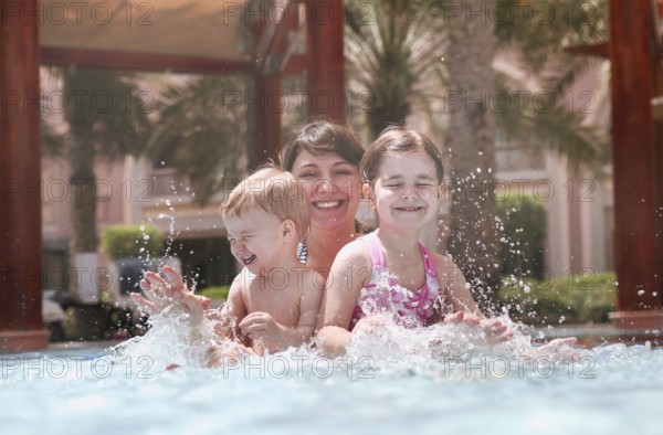 Overjoyed mother with little daughter and son laughing while playing together in swimming pool with on sunny day