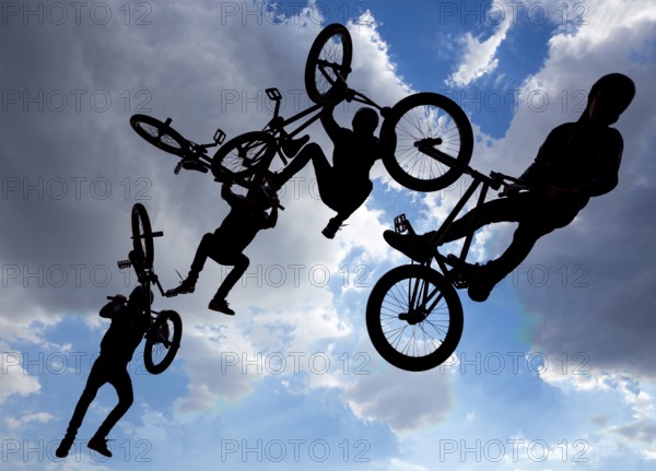 An extreme rider is making a free style jump from a ramp. The young boy with his bicycle is seen as a silhouette in front of the sun. Multiple exposure image. Clouds