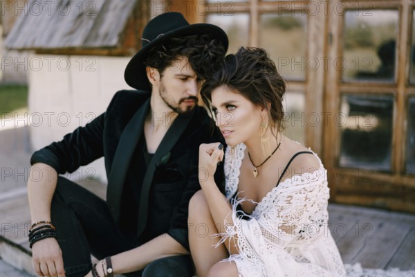 Groom in a black hat sits near bride in a wicker dress on the doorstep of a wooden house.