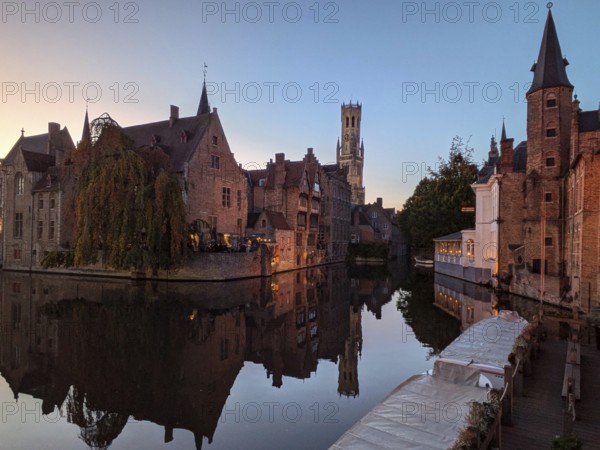 Charming canal at sunset with medieval buildings and a distant tower reflected in the calm water. Bruges, Belgium