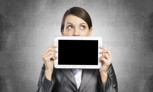 Businesswoman holding tablet computer with blank screen. Beautiful woman in business suit show tablet PC near her face. Corporate businessperson on grey wall background. Digital technology layout