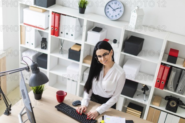 A young girl with glasses, wearing a white blouse and black trousers, works in the office. The girl has long straight black hair. photo with depth of field