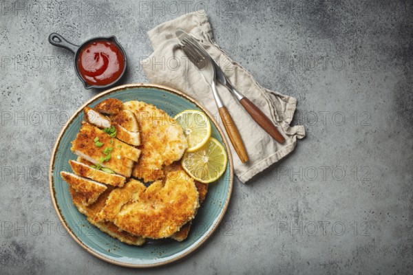 Crispy panko breaded fried chicken fillet with green salad and lemon cut on plate on gray background table with ketchup from above. Japanese style deep fried coated chicken breasts, space for text, food photography, food photography