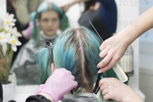 Back view of female head with emerald hair color and regrown hair roots. Woman sits in chair by mirror, two professional hairdressers combing client's hair before hair dyeing process in beauty salon