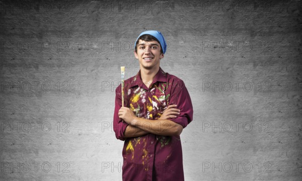 Young happy painter artist holding paintbrush. Portrait of painter in dirty shirt and bandana standing on gray wall background. Creative hobby and artistic occupation. Art school courses concept