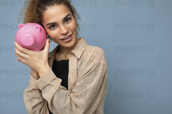 Portrait photo of happy positive cute young beautiful pretty brunette curly woman with sincere emotions wearing stylish beige shirt isolated over blue background with copy space and holding pink piggy box. Money box concept